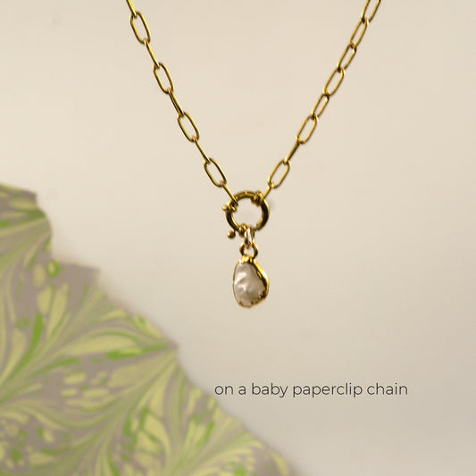 A gold paperclip chain necklace with a small gold clasp holding a irregular shaped white pearl pendant