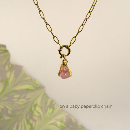 A gold paperclip chain necklace with a small gold ring clasp holding a rough Pink Tourmaline gemstone pendant,