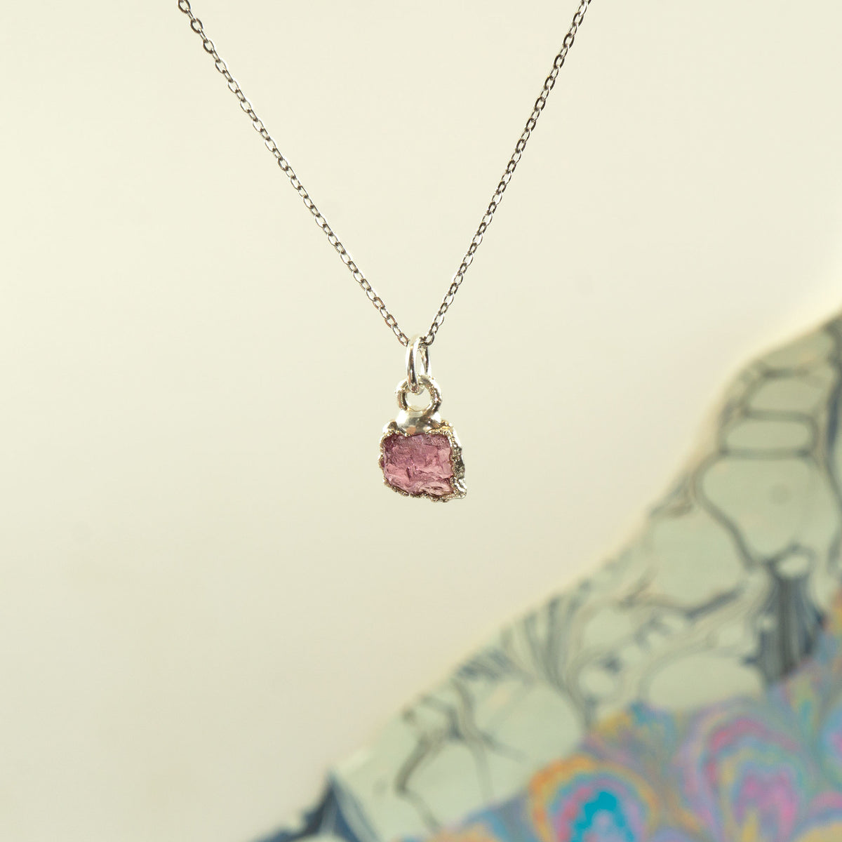 A delicate silver chain necklace with a small, rough-cut Pink Tourmaline gemstone pendant in a silver bezel setting,