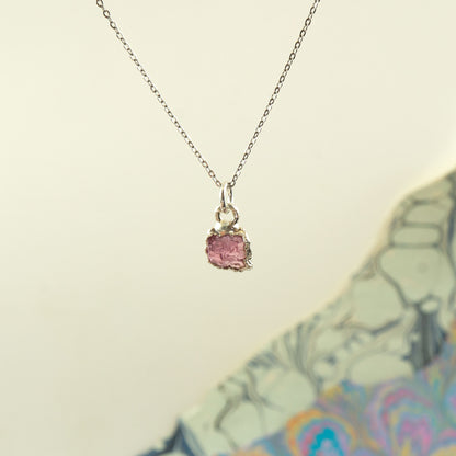 A delicate silver chain necklace with a small, rough-cut Pink Tourmaline gemstone pendant in a silver bezel setting,
