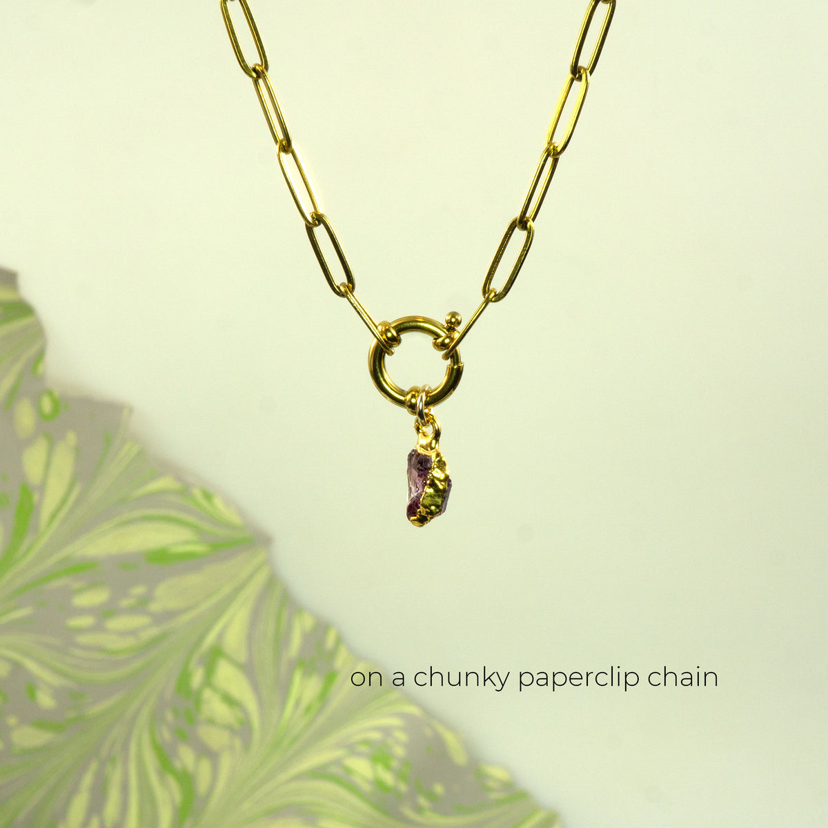 A gold paperclip chain necklace with a toggle clasp and a small dangling rhodlite garnet gemstone charm