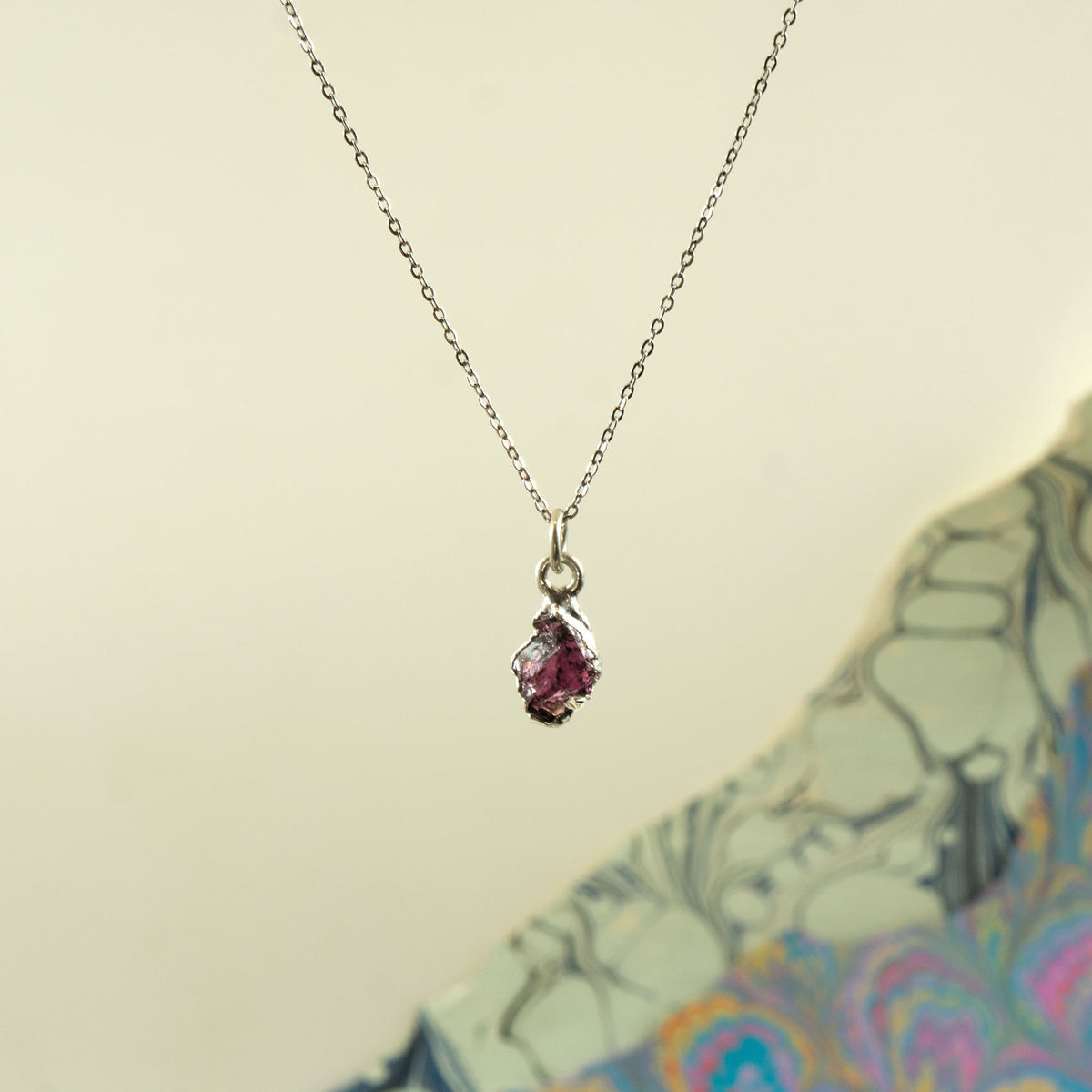 A delicate silver chain necklace with a small teardrop-shaped pendant featuring a pinkish-red rhodolite garnet gemstone