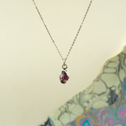 A delicate silver chain necklace with a small teardrop-shaped pendant featuring a pinkish-red rhodolite garnet gemstone