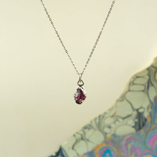 A delicate silver chain necklace with a small teardrop-shaped pendant featuring a pinkish-red rhodolite garnet gemstone