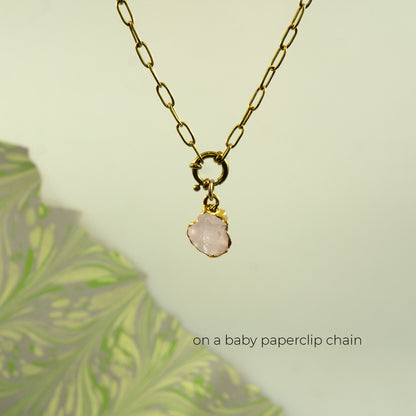 A gold paperclip chain necklace with a small gold clasp holding a light pink rose quartz stone pendant
