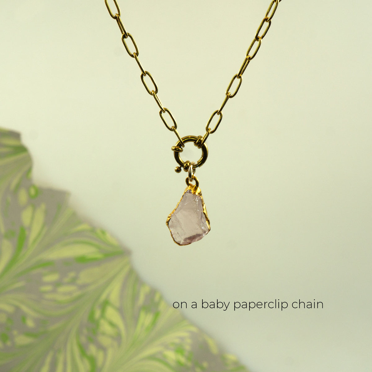 A gold paperclip chain necklace with a gold clasp holding a raw, irregular pale pink rose quartz stone pendant