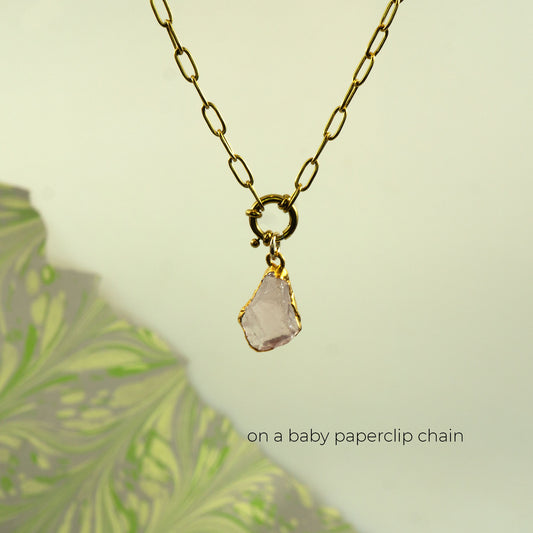 A gold paperclip chain necklace with a gold clasp holding a raw, irregular pale pink rose quartz stone pendant