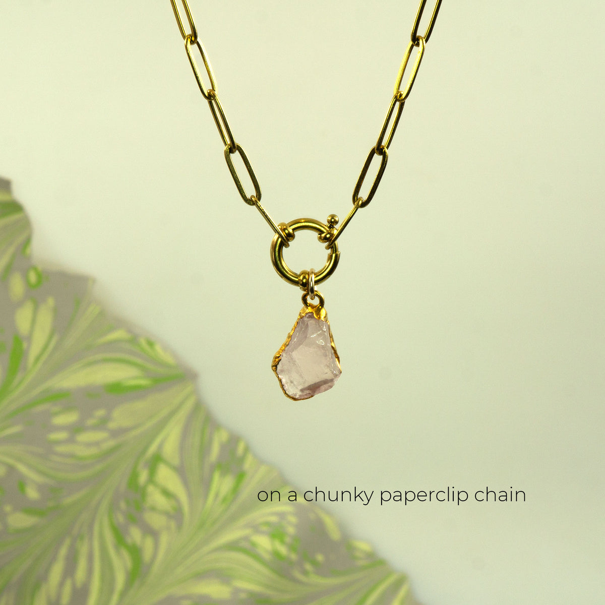 A gold paperclip chain necklace with a gold ring clasp and a raw, irregular pale pink rose quartz gemstone pendant