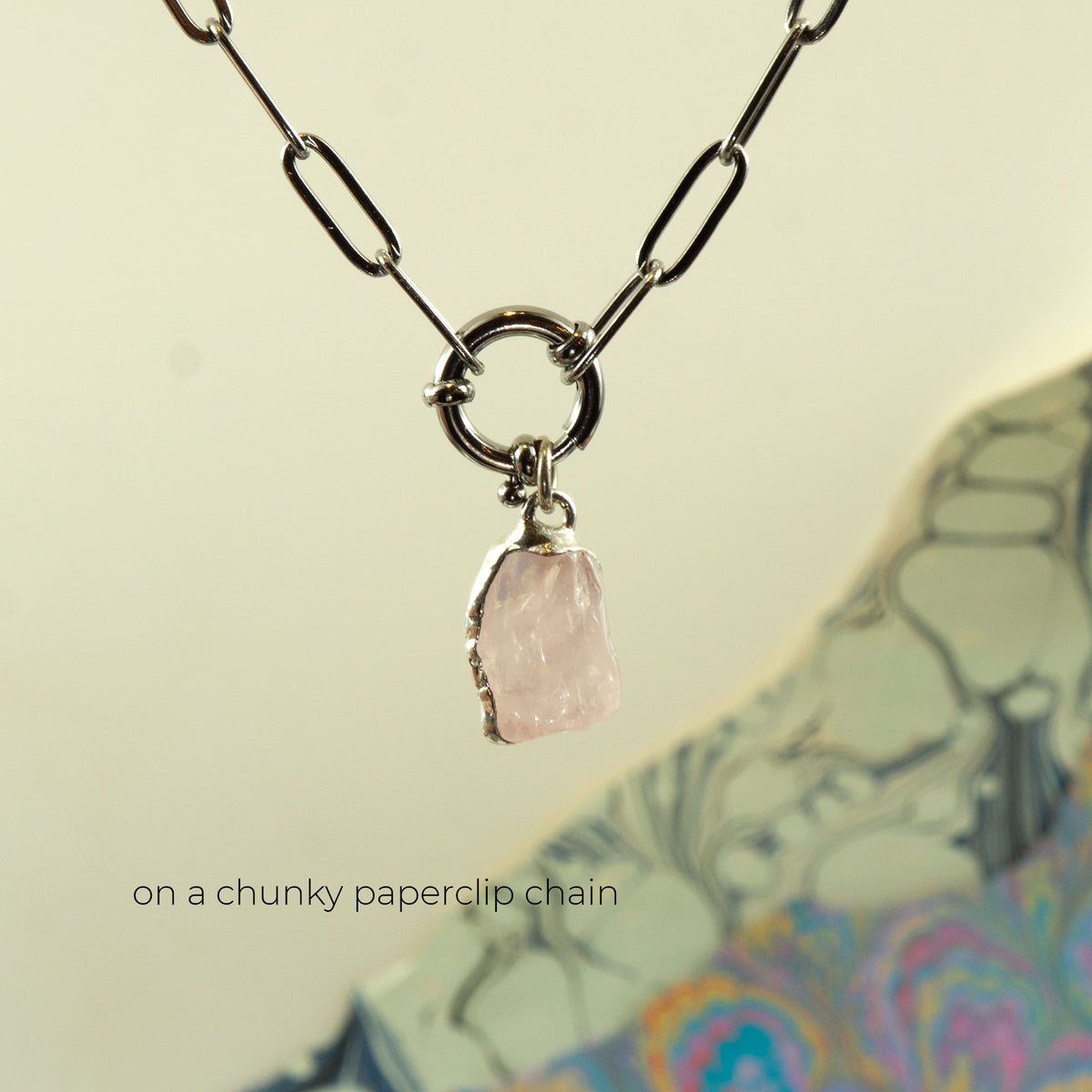 A silver paperclip chain necklace with a ring clasp holding a raw, irregular-cut pale pink raw rose quartz gemstone pendant