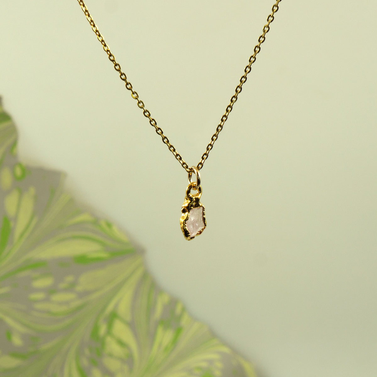 A delicate gold chain necklace with a small pendant featuring a rough, pale pink gemstone