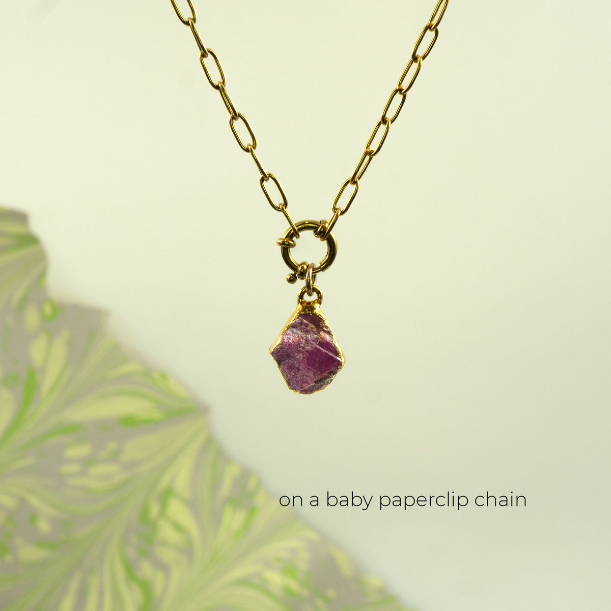 A gold chain necklace with a small gold ring clasp holding a rough, irregular ruby gemstone pendant