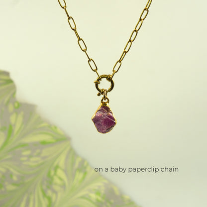 A gold chain necklace with a small gold ring clasp holding a rough, irregular ruby gemstone pendant