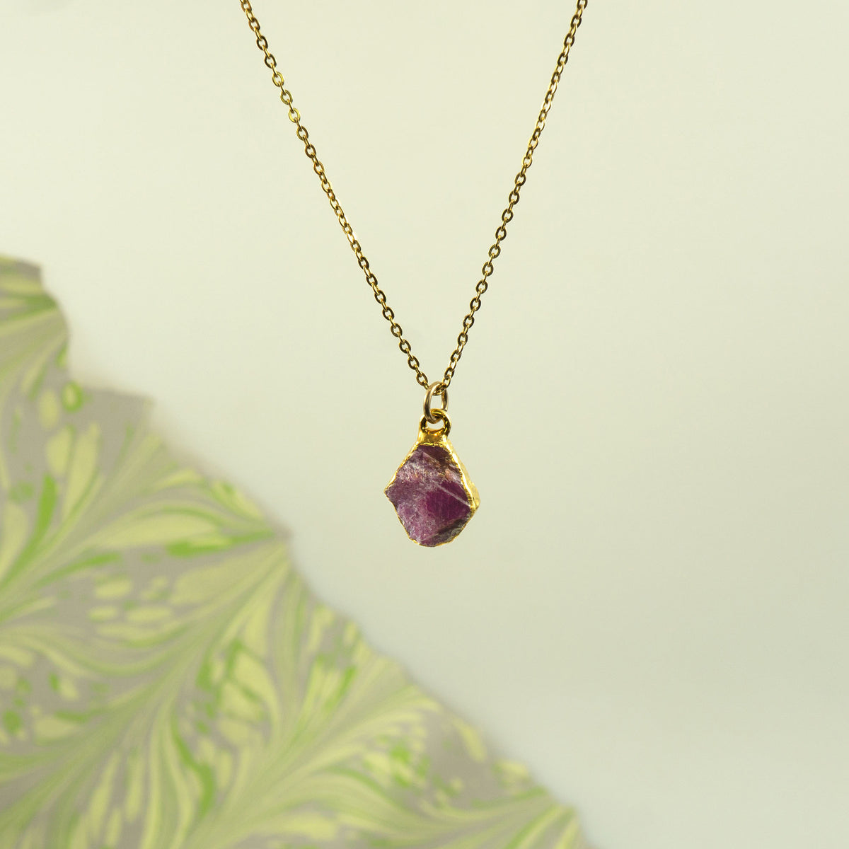 A gold chain necklace with a raw, irregular-cut ruby gemstone pendant