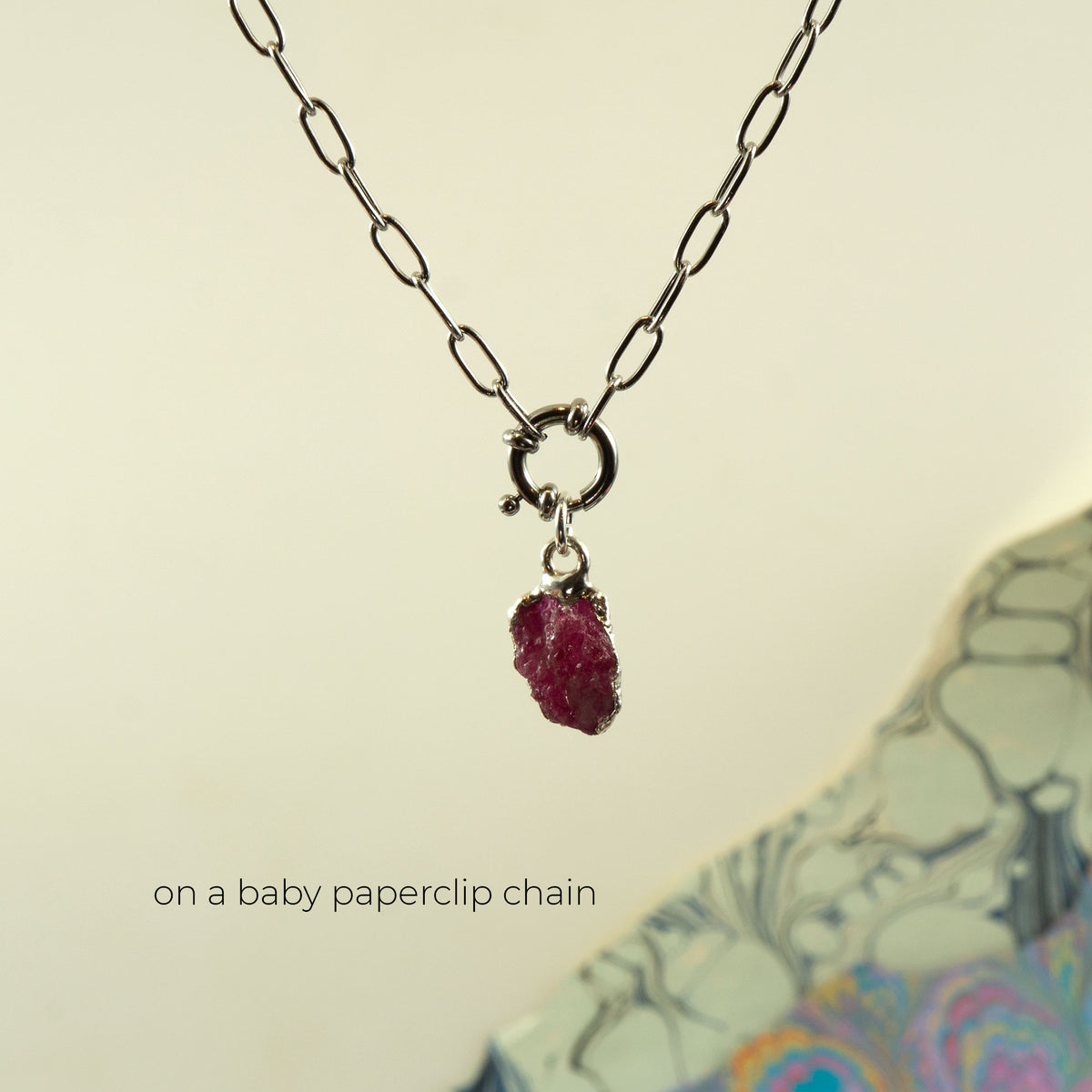 A silver paperclip chain necklace with a small ring clasp holding a raw, faceted ruby gemstone pendant