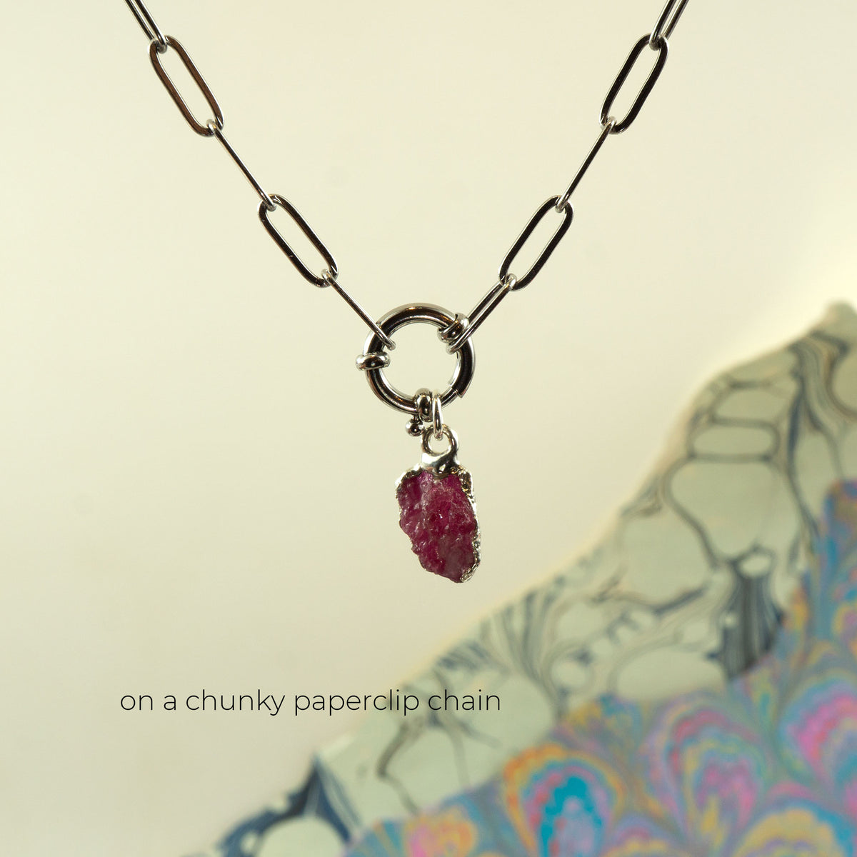 A silver paperclip chain necklace with a ring clasp holding a small raw ruby gemstone pendant,