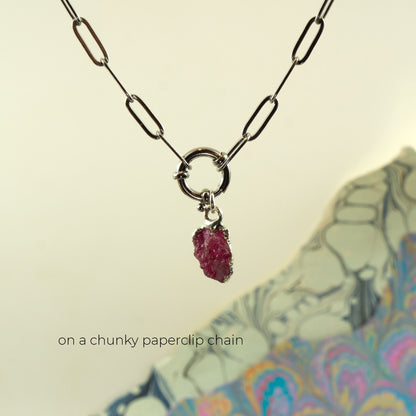 A silver paperclip chain necklace with a ring clasp holding a small raw ruby gemstone pendant,