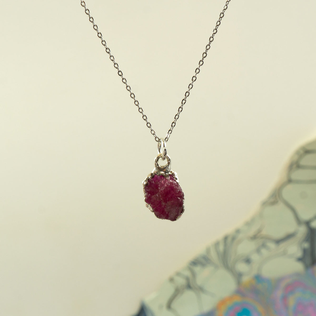 A silver chain necklace with a raw, rough ruby gemstone pendant