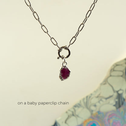 A silver paperclip chain necklace with a small ring clasp and a dangling raw ruby gemstone pendant