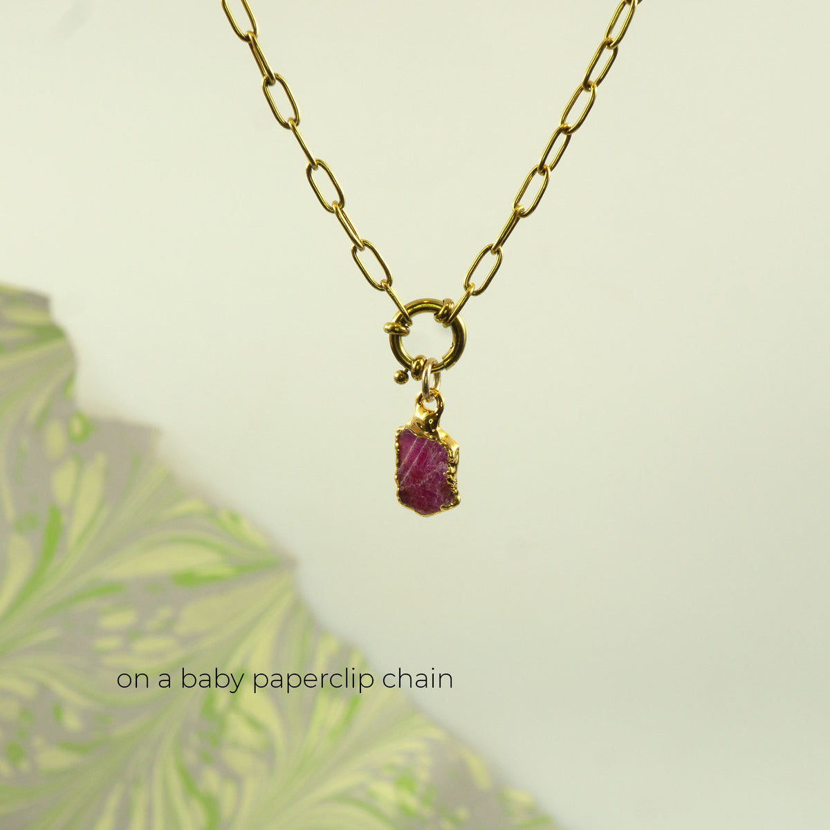 A gold paperclip chain necklace with a small gold clasp holding a rectangular ruby gemstone pendant