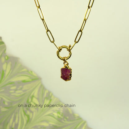 A gold paperclip chain necklace with a small ring connector and a raw, faceted ruby gemstone pendant