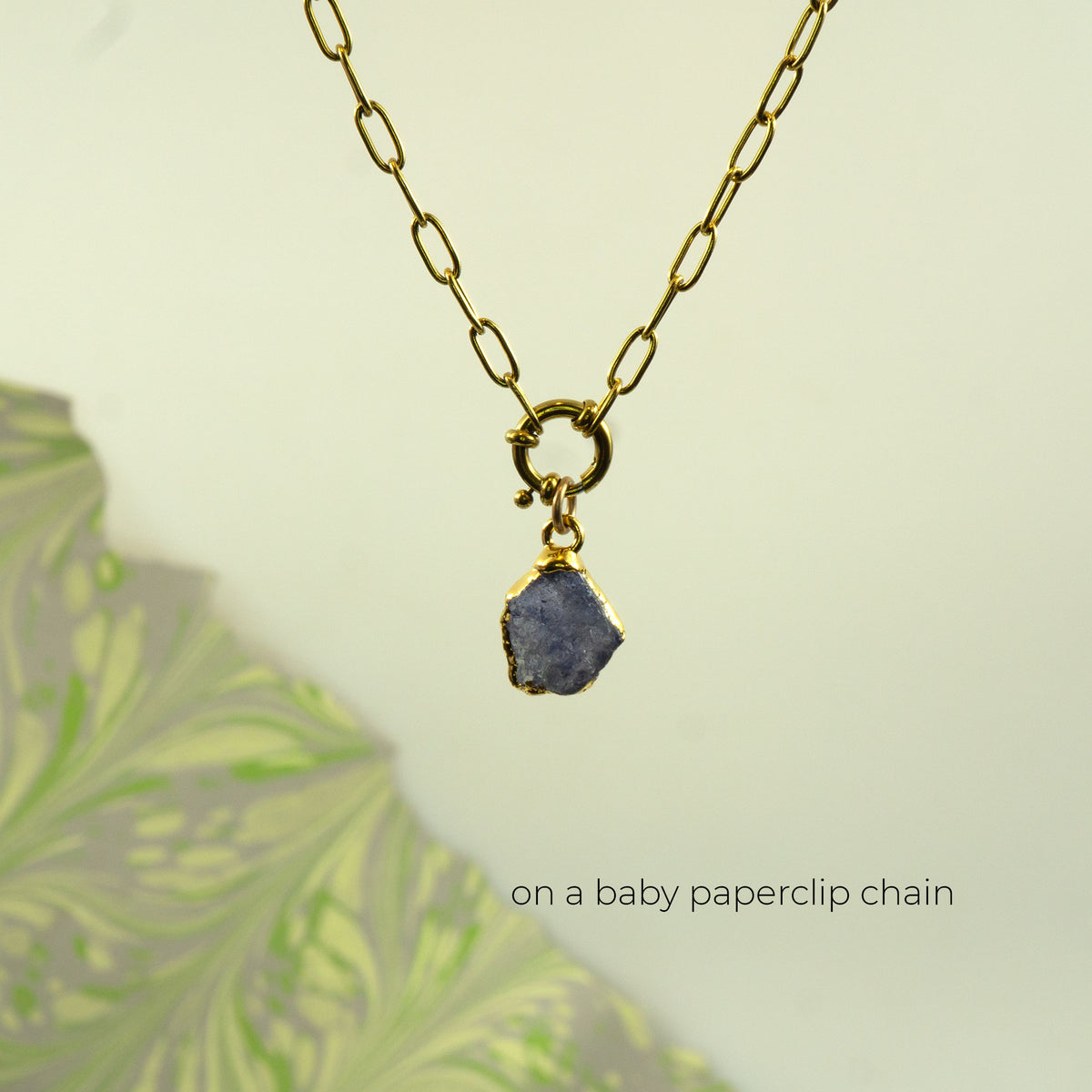A gold paperclip chain necklace with a small gold clasp holding a rough, irregular blue-gray tanzanite stone pendant