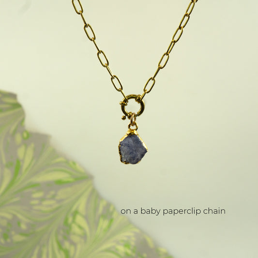 A gold paperclip chain necklace with a small gold clasp holding a rough, irregular blue-gray tanzanite stone pendant