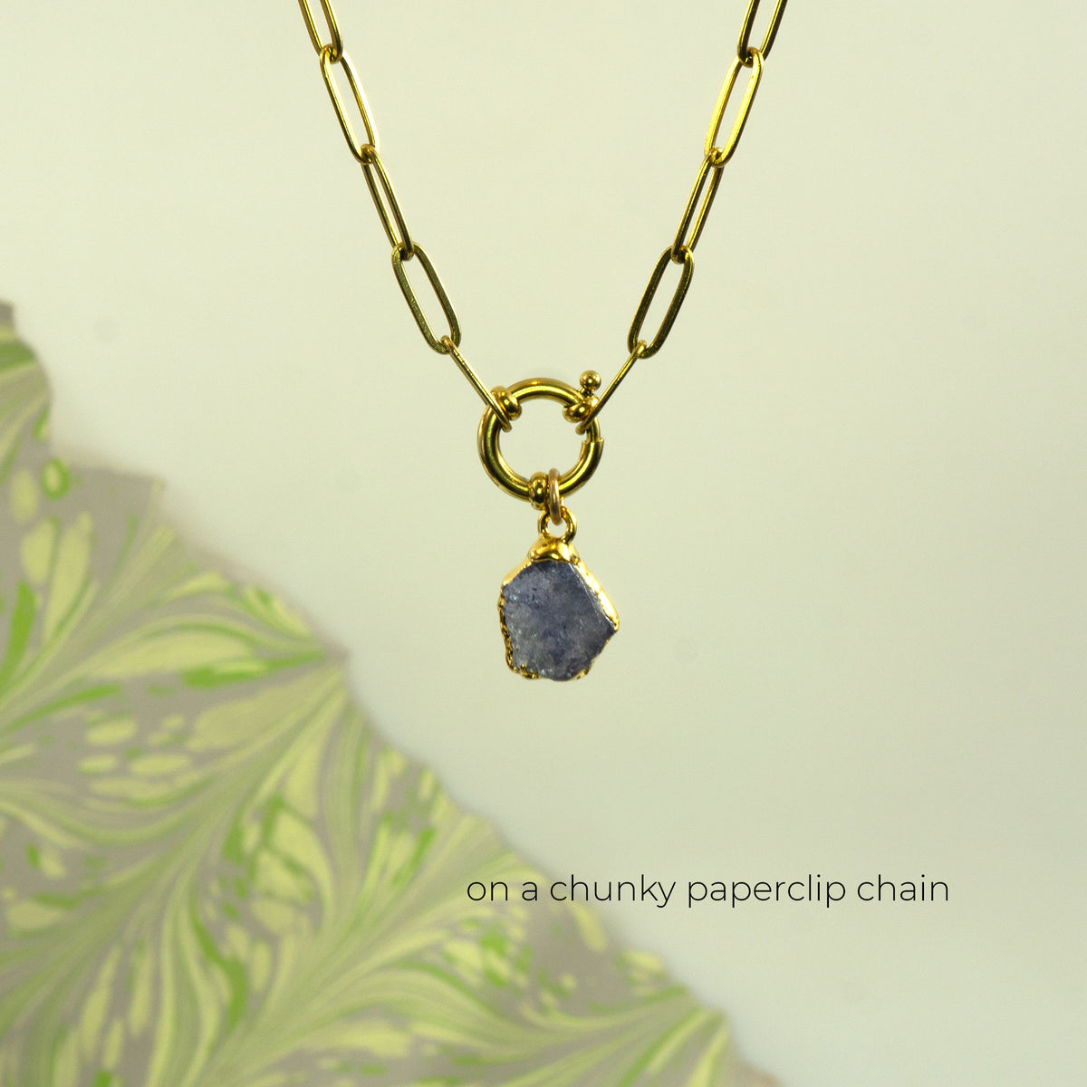 A gold paperclip chain necklace with a ring clasp holding a raw, irregular tanzanite gemstone pendant