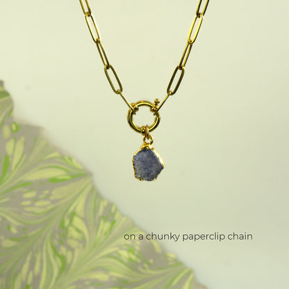 A gold paperclip chain necklace with a ring clasp holding a raw, irregular tanzanite gemstone pendant
