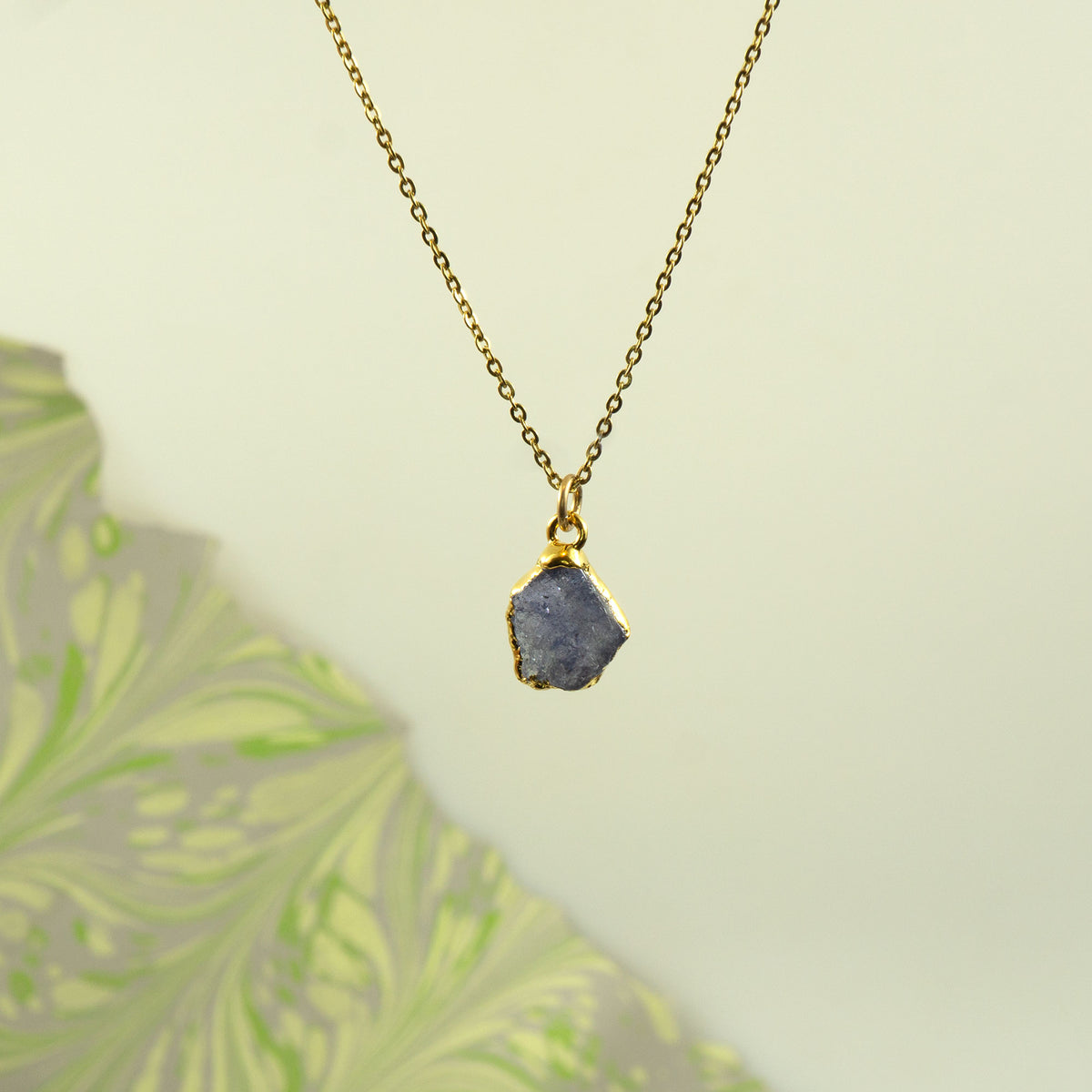 A gold chain necklace with a raw, irregular-shaped blue-gray tanzanite stone pendant