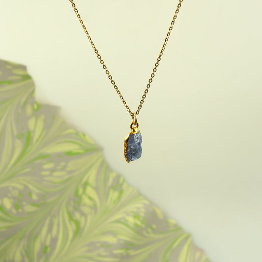 A gold chain necklace with a raw, irregular-shaped blue tanzanite stone pendant.