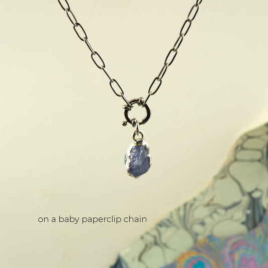 A silver paperclip chain necklace with a small ring clasp and a dangling raw, irregular blue tanzanite gemstone pendant