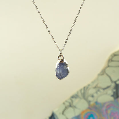 A silver chain necklace with a raw, irregular-shaped blue tanzanite gemstone pendant