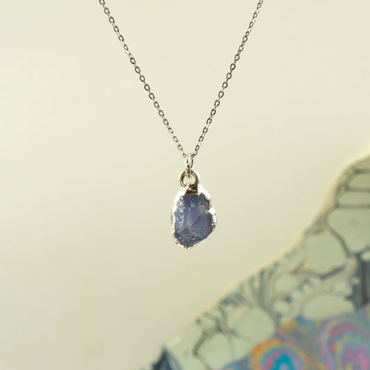 A silver chain necklace with a raw, irregular-shaped blue tanzanite gemstone pendant